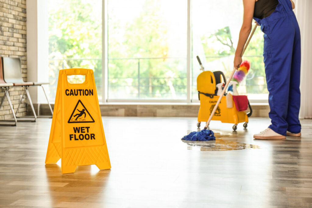 A person in blue overalls mops a wooden floor behind a bright yellow "CAUTION WET FLOOR" sign in a sunlit room.