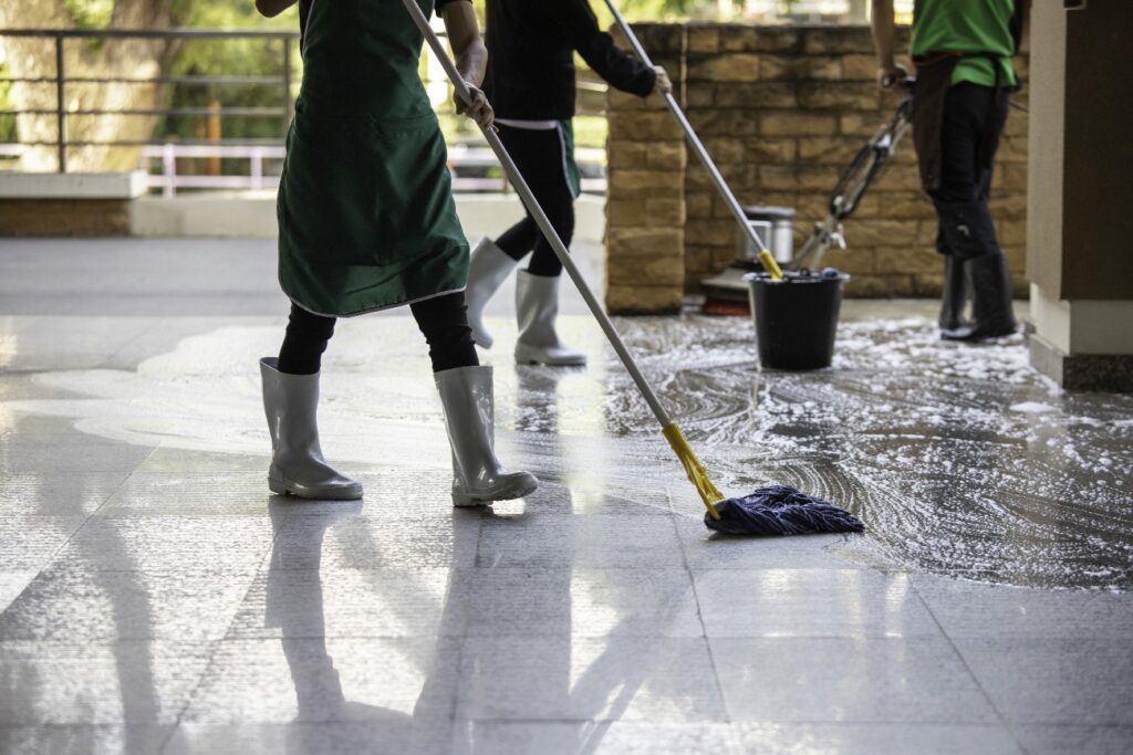 A team of cleaning staff in aprons and rubber boots scrubbing a wet, soapy tiled floor with mops and a floor buffer.