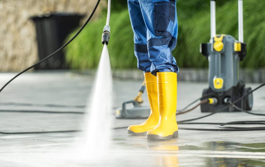 A person in blue overalls and yellow rubber boots operates a pressure washer to clean a surface outdoors.
